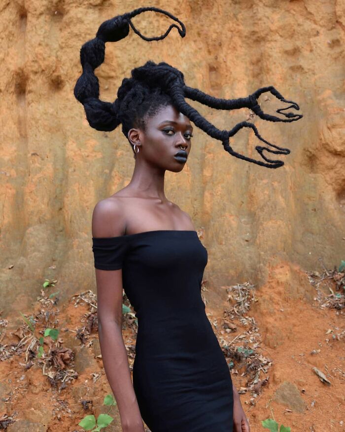Woman with hair sculpted into a scorpion shape, wearing an off-the-shoulder black dress, standing against a textured background.