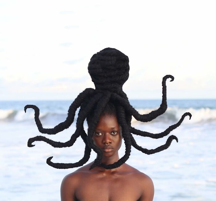 Artist with hair sculpture resembling an octopus, showcasing creativity against a beach backdrop.