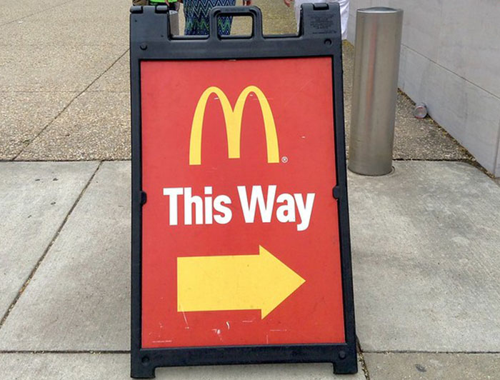 McDonald’s sidewalk sign with a yellow arrow pointing right, warning about chain restaurant employee advice.