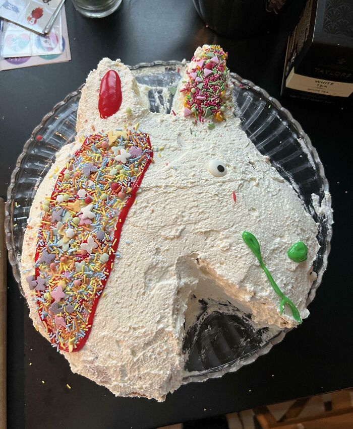 A unicorn-shaped cake with vibrant sprinkles and icing, partially eaten, a funny kitchen fail.