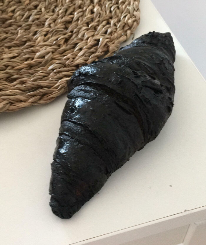 A charred croissant sitting on a white table next to a woven placemat, showing a funny cooking mishap.