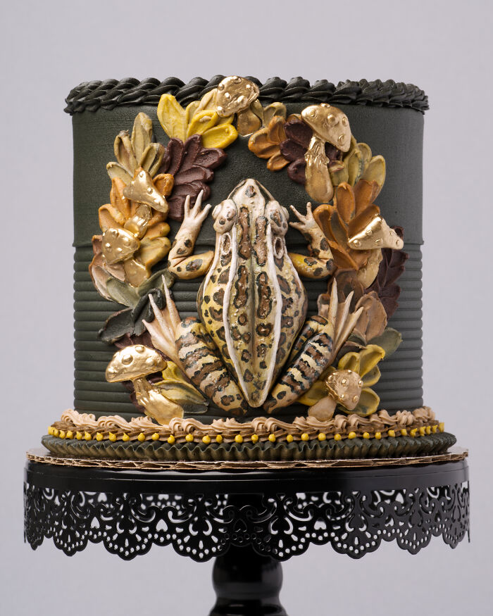 A whimsical vegan cake topped with a lifelike frog, nestled among edible leaves and mushrooms.