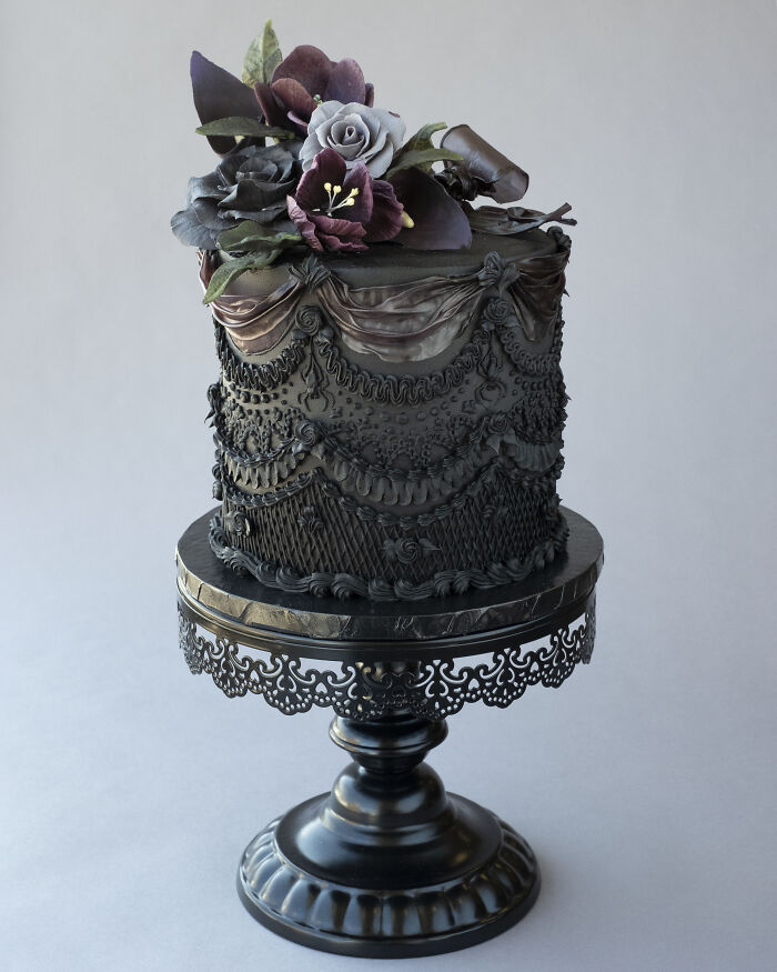 A delicate vegan cake draped in lacy icing with deep, dark flowers, perched on an elegant stand.